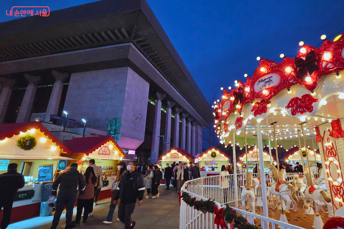 세종문화회관의 거대한 기둥이 보이는 광장 한쪽에 붉은 지붕의 먹거리 부스들이 줄지어 늘어서 있다. 오른쪽에는 화려한 조명의 회전목마가 일부 보이며, 많은 시민이 부스 사이를 오가며 축제를 즐기고 있다.