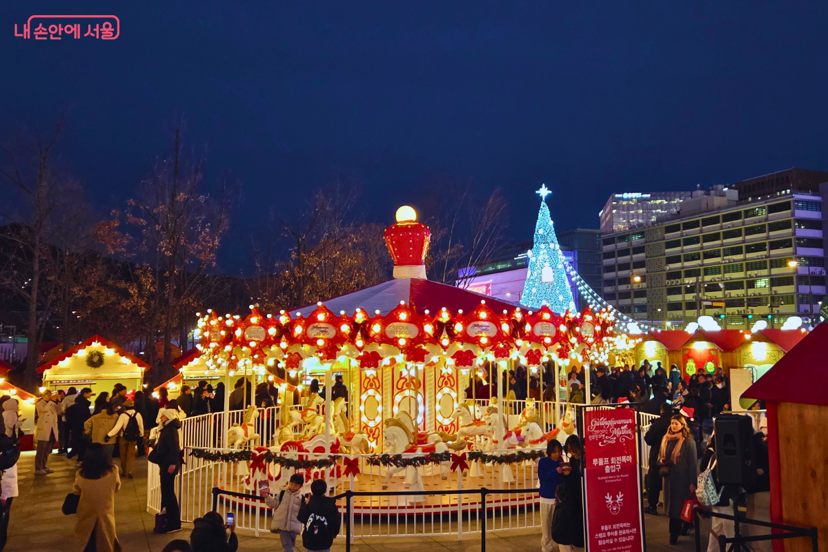 밤하늘 아래 화려하게 불을 밝힌 회전목마가 돌아가고 있다. 지붕은 붉은색과 흰색 줄무늬로 장식되었고, 붉은 리본 장식이 달려 있다. 시민들이 회전목마 주변에 모여 사진을 찍거나 구경하고 있으며, 뒤편으로 푸른색 대형 트리가 보인다.