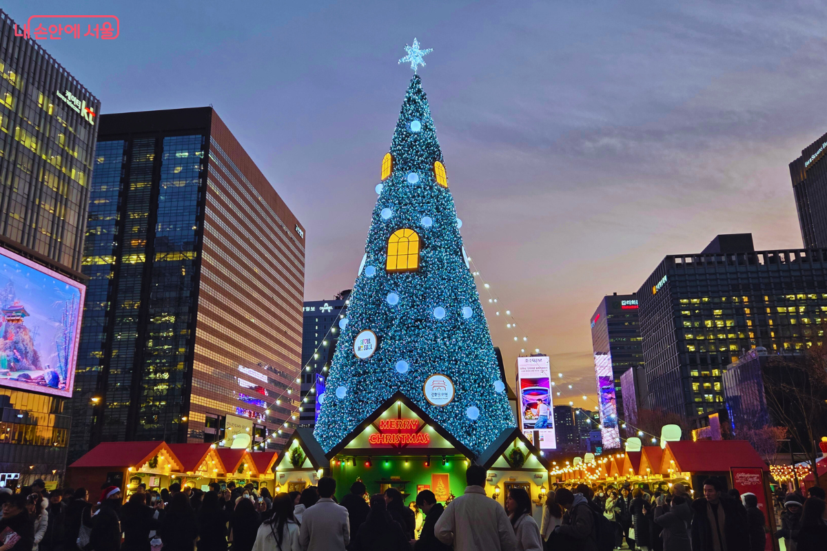 어둑해지는 저녁, 광화문광장에 설치된 거대한 푸른색 트리가 조명을 밝히고 있다. 트리 꼭대기에는 별 장식이 빛나며, 트리 주변과 마켓 부스 사이를 가득 메운 시민들이 연말 분위기를 즐기고 있다. 주변 고층 빌딩 숲과 어우러진 모습이다.