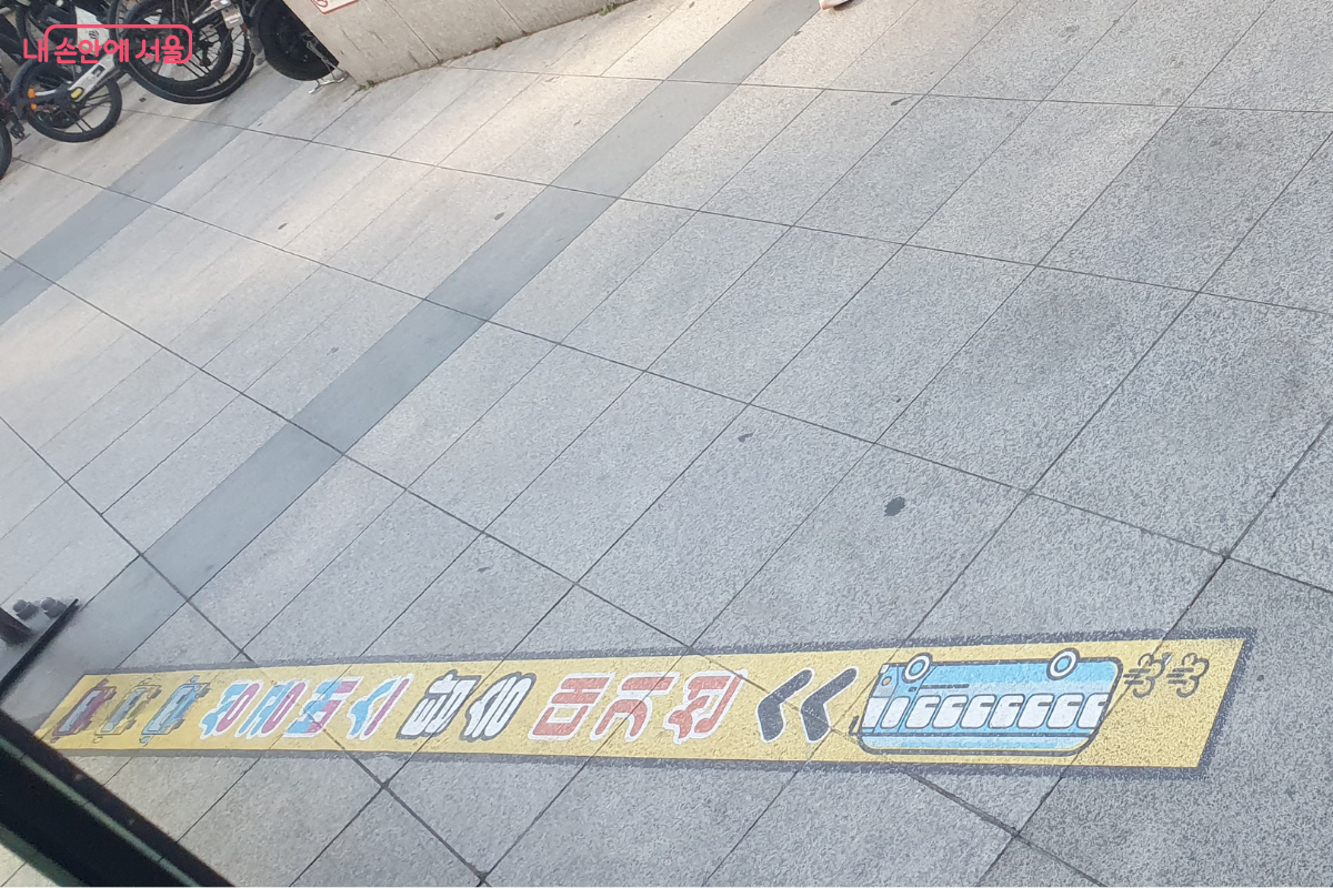 보도블록 바닥에 노란색 띠 형태로 부착된 '성공버스 탑승 대기선' 스티커이다. 귀여운 버스 일러스트와 함께 화살표 방향으로 줄을 서 달라는 안내가 되어 있어, 시민들이 질서 있고 안전하게 버스를 기다릴 수 있도록 유도한다. ©양정화