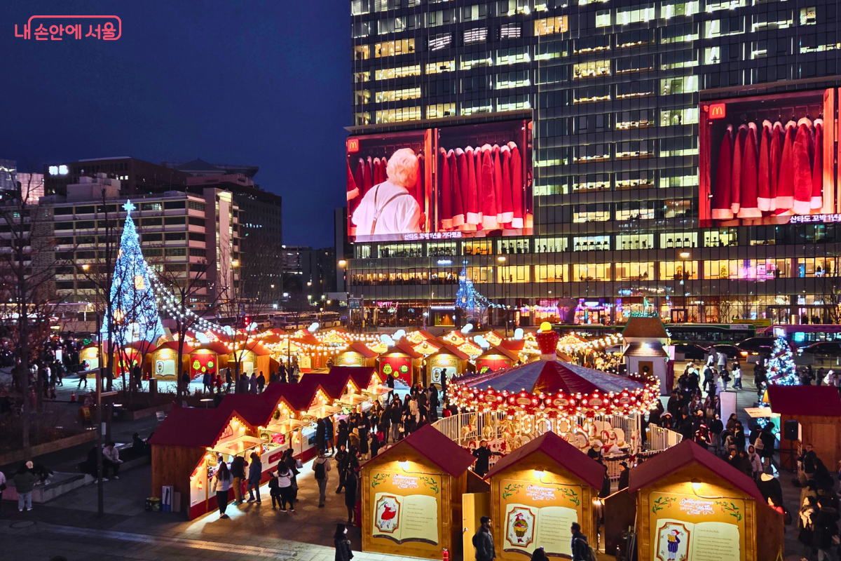 높은 곳에서 내려다본 광화문광장의 야경이다. 왼쪽에는 대형 트리가, 오른쪽에는 회전목마가 빛나고 있다. 뒤편 건물 대형 전광판에는 산타클로스 복장이 걸린 옷장 영상이 나오고 있어 크리스마스 분위기를 더한다.