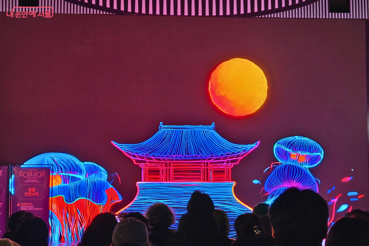 야외 스크린에 미디어아트가 상영되고 있다. 붉은색과 푸른색 네온 선으로 광화문 누각과 둥근 보름달, 해파리 같은 형상이 그려져 있다. 화면 앞에는 많은 시민이 모여 작품을 감상하고 있다.