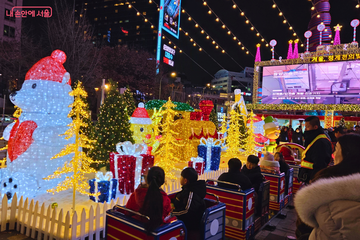 청계광장에 설치된 크리스마스 테마의 어린이 놀이 기구인 '어린이 트레인'의 모습. 빨간색 기차 모양의 좌석에 아이들이 탑승하여 즐거워하고 있다. 주변에는 흰색 곰 모양의 대형 조형물과 노란색 빛의 미니 트리, 그리고 산타 모자를 쓴 눈사람 모양의 조형물들이 장식되어 있다. 배경의 대형 전광판은 밝은 보라색 빛을 내며 축제의 활기찬 분위기를 더한다.