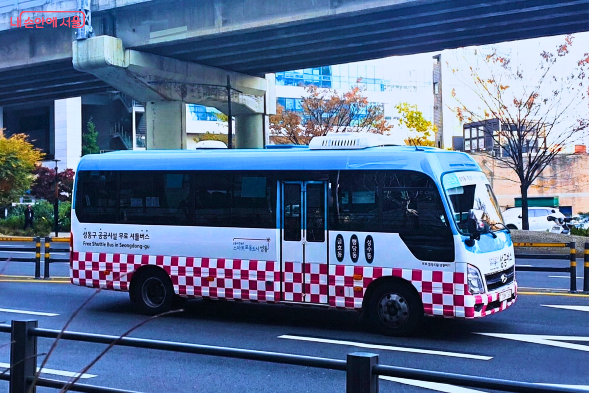 파란색과 흰색, 체크무늬 디자인이 돋보이는 성동구 '성공버스'가 도로를 달리고 있다. 버스 측면에는 '성동구 공공시설 무료 셔틀버스'라는 문구와 함께 금호동, 행당동, 성수동 등 주요 경유지 배지 이미지가 부착되어 있어, 지역 주민을 위한 교통 복지 수단임을 보여준다. ©양정화