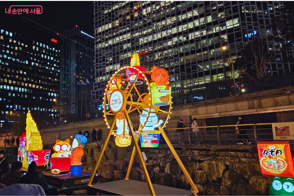 청계천 4구역으로 추정되는 곳에 설치된 대형 관람차 모양의 유등 조형물. 관람차 각 칸에는 농심의 대표 캐릭터들이 라면 등 자사 제품을 들고 앉아 있는 모습이 표현되어 있다. 관람차는 노란색과 주황색을 주 색상으로 하고 있으며, 주변에는 노란색 미니 트리와 다른 캐릭터 유등이 함께 배치되어 있다. 뒤쪽으로는 고층 빌딩들이 밤하늘을 배경으로 하고 있다.