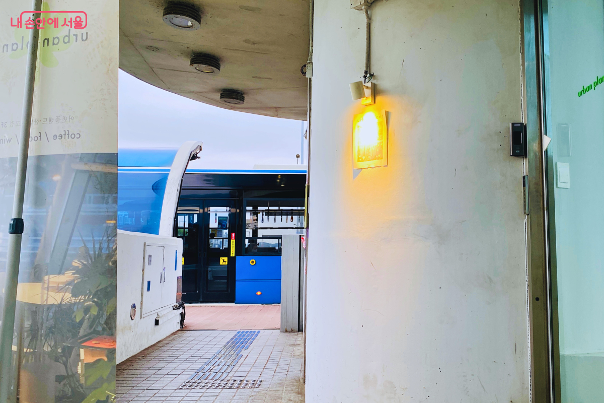 카페 입구의 하얀 벽면 뒤로 버스정류장에 정차한 파란색 간선버스의 뒷모습이 보인다. 입구 벽에는 따뜻한 오렌지빛 조명이 켜져 있으며, 바닥은 격자무늬 타일로 마감되어 있다. 버스 정류장이 카페와 얼마나 밀접해 있는지 보여준다. &copy;양정화