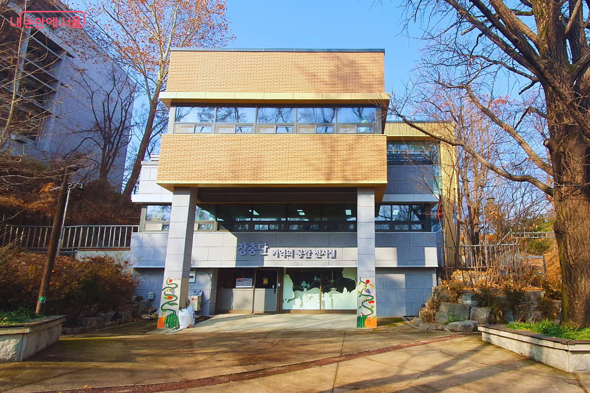 '장충단, 기억의 공간 전시실' 건물의 정면 외관이다. 현대적인 감각의 2층 석조 건물로, 1층 입구 기둥에는 초록색 식물 넝쿨 그림이 장식되어 있다. 건물 정면에는 전시실 명칭이 뚜렷하게 부착되어 있으며, 주변의 커다란 활엽수들이 건물을 감싸고 있어 차분하고 정적인 느낌을 준다. &copy;양정화