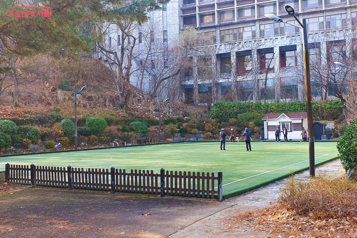 장충단공원 한쪽에 마련된 푸른 인조 잔디 게이트볼 경기장이다. 낮은 나무 울타리로 구획된 경기장에서 시민들이 게이트볼 경기를 즐기고 있으며, 뒤편으로는 작은 목조 대기소와 담쟁이덩굴로 덮인 현대식 건물이 보인다. 공원의 자연경관과 어우러져 평화로운 여가 활동의 현장을 보여준다. &copy;양정화