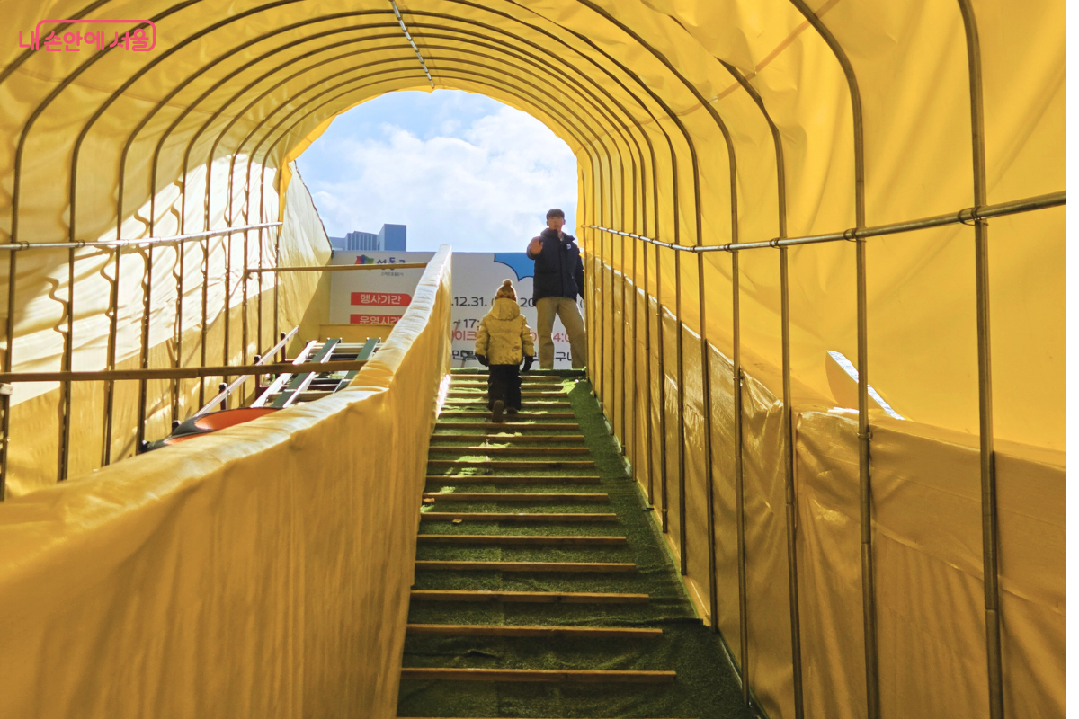 노란색 천막으로 덮인 터널형 슬로프 이동 통로 내부의 모습이다. 중앙에는 아이들이 걸어 올라갈 수 있도록 인조 잔디가 깔린 계단형 통로가 있고, 왼쪽에는 무거운 튜브를 자동으로 운송해 주는 이동식 컨베이어 벨트 설비가 가동되고 있다. 통로 끝 정상부에는 안전요원이 서서 이용객들을 맞이하고 있으며, 천막 사이로 들어오는 햇살이 내부를 환하게 비추고 있다. &copy;양정화