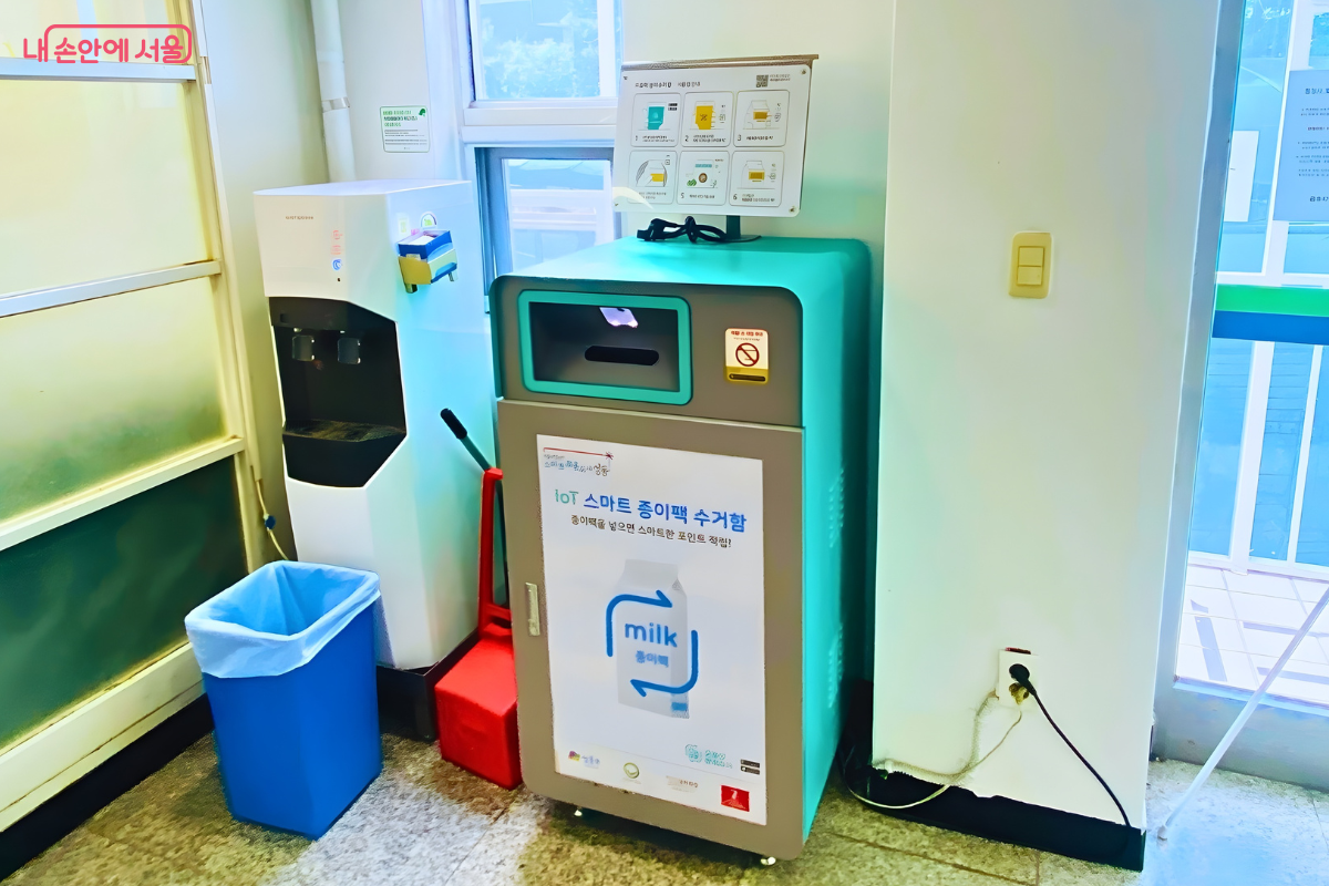 금호4가동 주민센터 실내 한쪽 벽면에 설치된 청록색과 회색 조화의 IoT 스마트 종이팩 수거함 모습이다. 수거함 앞면에는 '종이팩을 넣으면 스마트한 포인트 적립'이라는 문구와 우유팩 일러스트가 크게 부착되어 있으며, 상단에는 상세 사용법 안내판이 거치되어 있다. 수거함 왼쪽에는 파란색 분리수거함과 빨간색 손수레가 놓여 있고, 오른쪽 벽면에는 전원 코드가 연결된 상태이다.