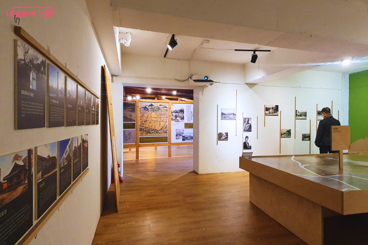 장충단, 기억의 공간 전시실 내부에서 한 시민이 전시물을 관람하고 있는 모습이다. 흰색 벽면을 따라 장충단과 관련된 인물, 사건의 사진 자료들이 일정한 간격으로 전시되어 있으며 상단 조명이 각 작품을 비추고 있다. 중앙에는 입체적인 지형 모델이 설치되어 있어 당시의 공간적 배경을 이해하도록 돕는다. &copy;양정화