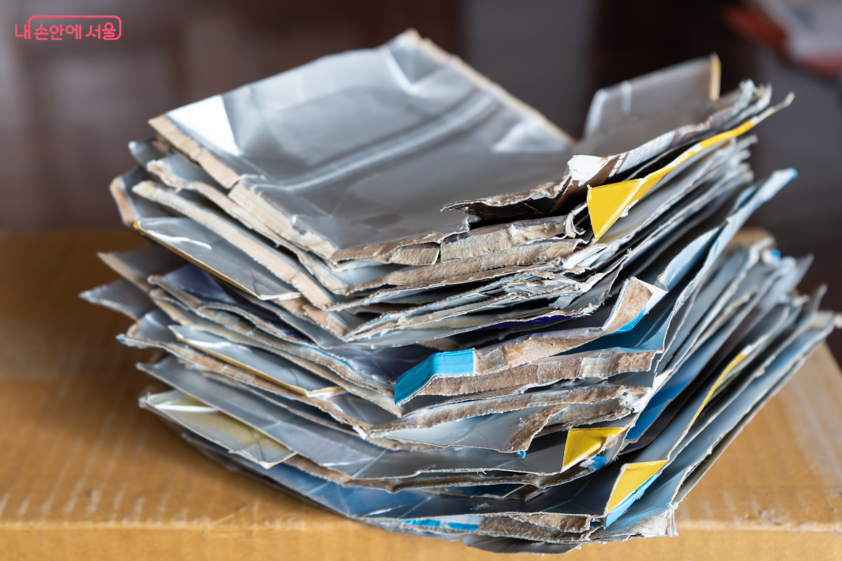 갈색 박스 위에 납작하게 접혀서 겹겹이 쌓여 있는 종이팩들의 상세 모습이다. 종이팩의 찢어진 단면을 통해 내부가 알루미늄으로 코팅된 멸균팩 재질임을 육안으로 확인할 수 있다. 이러한 코팅 재질 때문에 일반 폐지와 섞일 경우 재활용이 어려우며 반드시 전용 수거함에 배출해야 함을 보여준다.