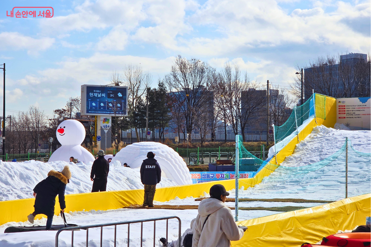 하얀 눈이 가득한 눈놀이터의 모습이다. 화면 중앙에는 '스태프(STAFF)' 문구가 적힌 검은색 방한복을 입은 안전요원이 뒷모습을 보이며 서서 현장을 살피고 있다. 왼쪽에는 빨간 코가 달린 대형 눈사람 에어 인형과 이글루 모형이 보이며, 배경의 전광판에는 '현재날씨 3℃'와 기상 정보가 표시되고 있다. 아이들과 시민들이 안전요원의 관리 아래 자유롭게 눈놀이를 즐기는 평화로운 풍경이다. &copy;양정화