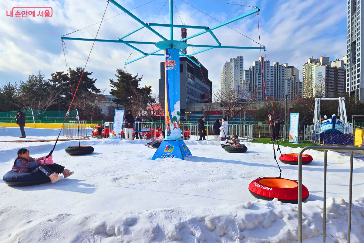 썰매장 한가운데 설치된 회전썰매의 전경이다. 파란색 기둥을 중심으로 여러 개의 줄이 연결되어 있으며, 줄 끝에 달린 튜브 썰매에 아이들이 앉아 눈 위를 빙글빙글 돌며 즐거워하고 있다. 기둥에는 '무지개 눈썰매장' 로고가 새겨진 현수막이 감겨 있고, 바닥은 하얀 눈으로 가득하다. 주변으로는 미니 기차 선로와 도심 건물들이 어우러져 활기찬 축제 분위기를 자아낸다. &copy;양정화