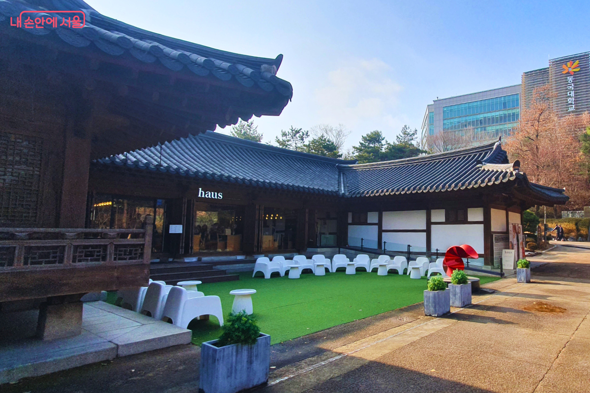 장충단공원 내 위치한 한옥 카페 '하우스(haus)'의 전경이다. 기와지붕 아래 현대적인 유리창이 조화를 이루는 건물 앞으로 푸른 인조 잔디가 깔린 마당이 펼쳐져 있으며, 흰색 디자인 의자들이 놓여 있다. 마당 한쪽에는 붉은색 조형물이 포인트로 설치되어 있어 세련된 휴식 공간의 분위기를 자아낸다. &copy;양정화