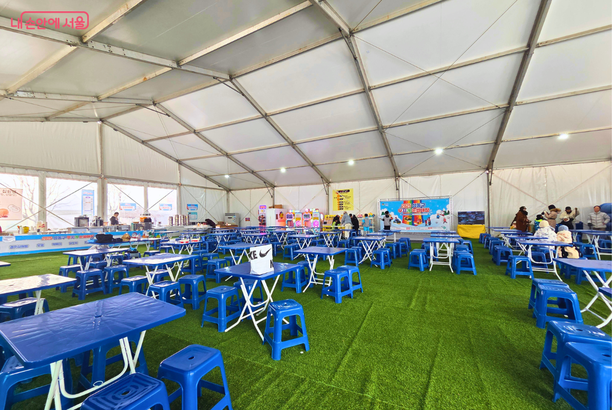 넓은 실내 휴게공간의 전체 전경이다. 바닥에는 초록색 인조 잔디가 깔려 있고, 수십 개의 파란색 간이 테이블과 의자가 일정한 간격으로 질서 정연하게 배치되어 있다. 천막 천장 사이로 밝은 채광이 들어와 내부가 환하며, 왼쪽 벽면에는 분식을 판매하는 매점 구역이 길게 마련되어 있다. 벽면 곳곳에는 이용 안내문과 메뉴판이 부착되어 시민들이 편리하게 이용할 수 있는 구조다. &copy;양정화