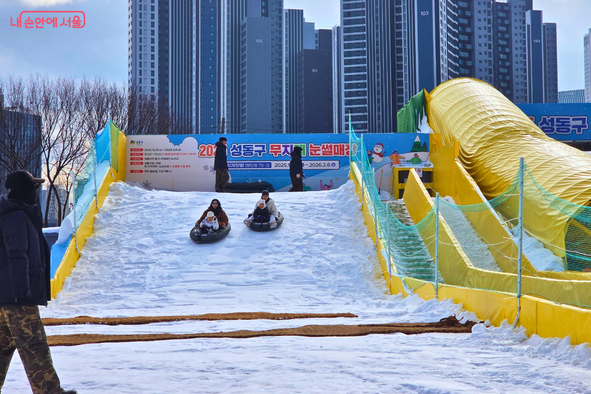 영유아용 슬로프에서 엄마와 아이가 함께 2인용 튜브 썰매를 타고 내려오는 모습이다. 두 가족이 각각 커다란 검은색 튜브에 앉아 하얀 눈 위를 미끄러져 내려오고 있으며, 아이들은 보호자의 품에 안겨 안전하게 썰매를 즐기고 있다. 슬로프 양옆은 노란색 가드와 초록색 그물망으로 안전하게 보호되어 있으며, 뒤쪽으로는 고층 아파트 건물들이 도심의 풍경을 이루고 있다. &copy;양정화