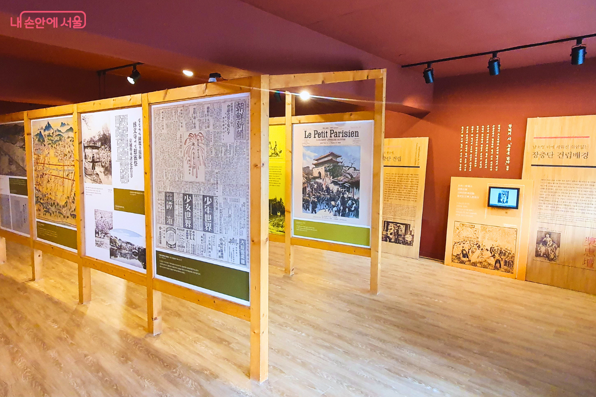 전시실의 또 다른 전시 구역으로, 나무 프레임을 활용해 과거의 신문 기사와 삽화들을 입체적으로 배치한 공간이다. 붉은 빛이 도는 벽면에는 장충단 건립 배경에 대한 상세한 설명글이 적혀 있고, 오른쪽 하단 모니터에서는 관련 영상이 재생되고 있어 관람객의 이해를 돕는다. &copy;양정화