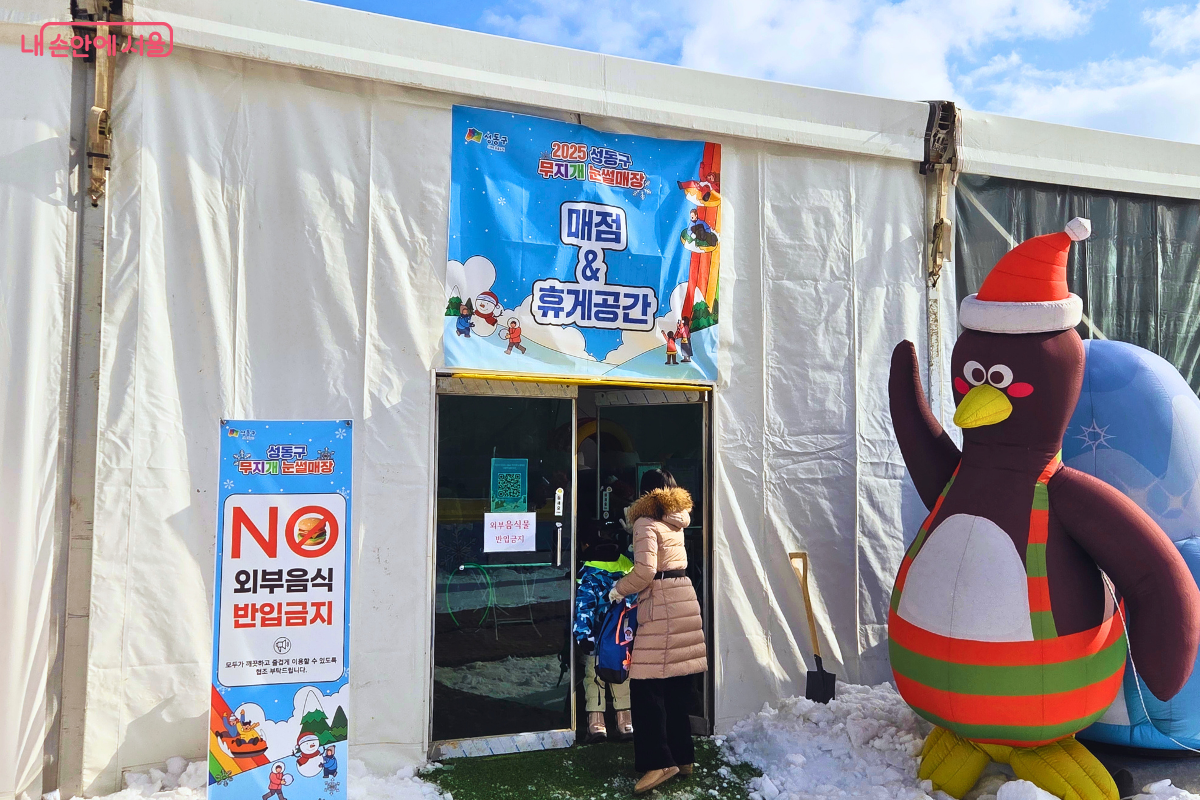 대형 하얀색 천막으로 조성된 실내 편의시설의 입구 모습이다. 문 위에는 '매점 & 휴게공간'이라고 적힌 현수막이 걸려 있고, 왼쪽에는 'NO 외부음식 반입금지'라고 적힌 파란색 안내 배너가 세워져 있다. 입구 오른쪽에는 산타 모자를 쓴 커다란 펭귄 에어 인형이 손을 흔드는 포즈로 서 있으며, 두꺼운 외투를 입은 시민들이 천막 안으로 입장하고 있다. &copy;양정화