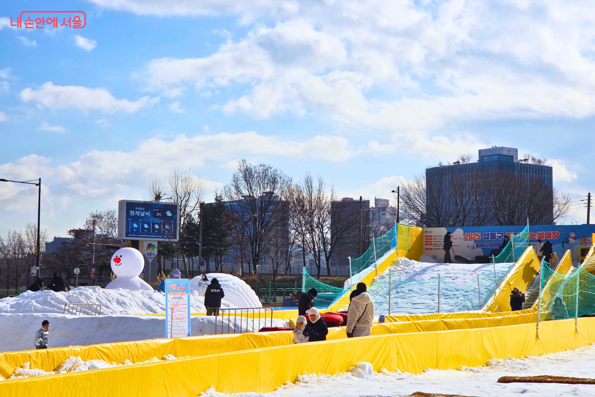 맑은 겨울 하늘 아래 조성된 영유아 전용 슬로프와 눈놀이동산의 풍경이다. 왼쪽에는 커다란 하얀색 눈사람 에어 인형이 세워져 있고, 그 옆으로는 아이들이 눈싸움을 하거나 눈사람을 만들며 놀 수 있는 눈놀이동산이 넓게 펼쳐져 있다. 오른쪽에는 경사가 완만한 영유아용 슬로프가 있으며, 노란색 안전 펜스가 설치되어 슬로프 구역을 안전하게 구분하고 있다. &copy;양정화