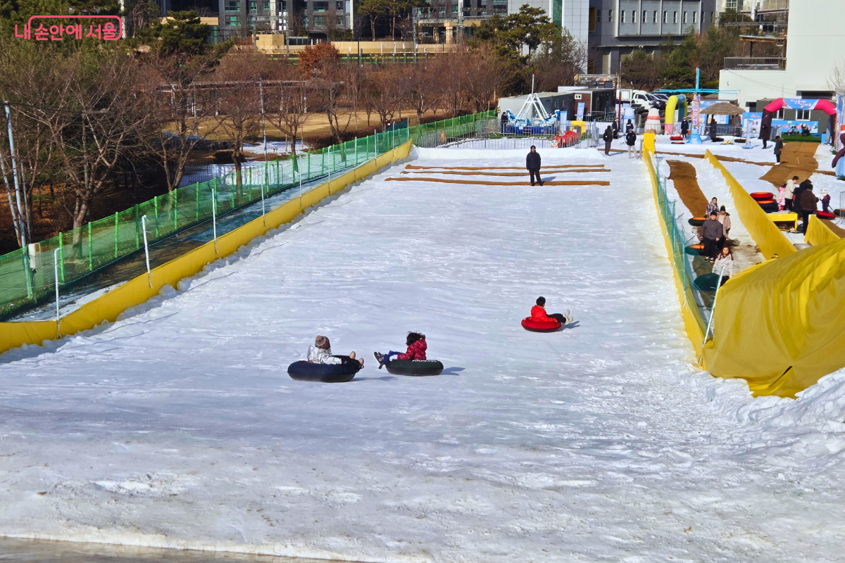 대형 슬로프에서 이용객들이 튜브 썰매를 타고 시원하게 내려가는 현장 사진이다. 하얀 눈 위로 세 명의 이용객이 각각 튜브에 앉아 서로 다른 지점을 미끄러져 내려가고 있으며, 슬로프 바닥에는 속도 조절을 위한 갈색 패드가 중간중간 설치되어 있다. 오른쪽에는 노란색 터널 통로가 길게 이어져 있고, 배경으로는 겨울 나무들과 도심의 건물들이 조화를 이루고 있다. &copy;양정화