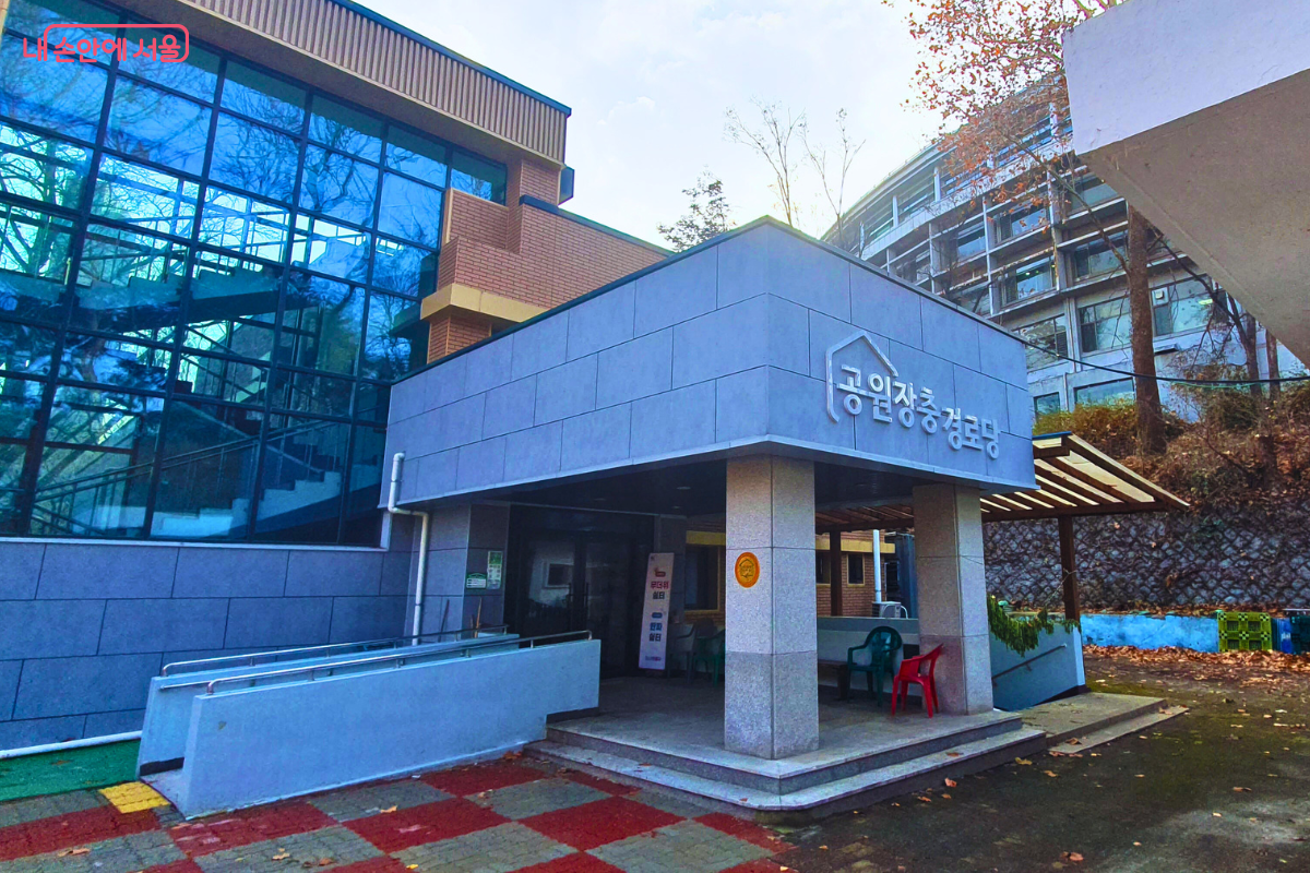 장충단공원 내부에 위치한 '공원장충경로당' 건물의 전경이다. 회색 석재와 통유리로 마감된 현대식 건물로, 입구에는 무더위 및 한파 쉼터를 알리는 표지판이 세워져 있다. 건물 왼쪽으로는 내부 계단이 보이는 유리 통창 구조가 특징적이며, 어르신들을 위한 쾌적한 휴식 공간임을 알 수 있다. &copy;양정화