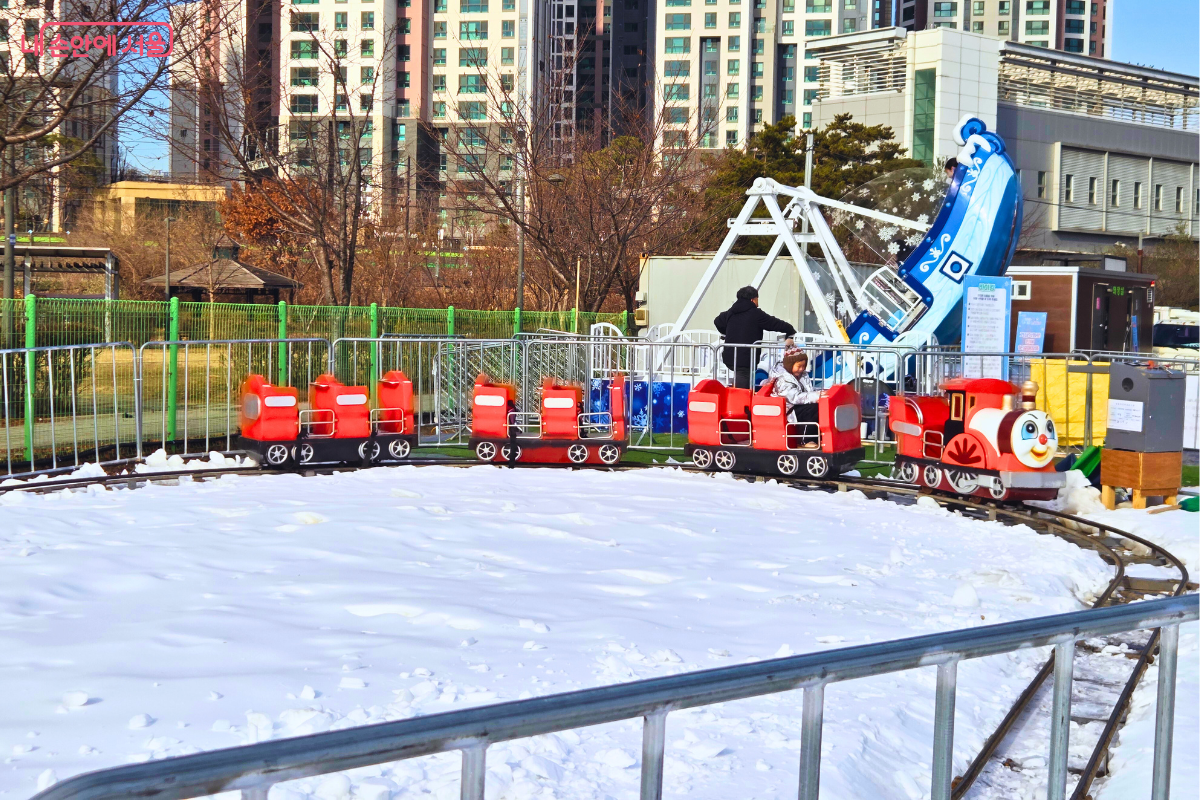 하얀 눈이 덮인 원형 선로 위를 빨간색 미니 기차가 달리고 있는 모습이다. 기차의 맨 앞칸에는 눈 큰 캐릭터 얼굴이 그려져 있으며, 칸마다 어린이가 한 명씩 앉아 즐겁게 기차 여행을 만끽하고 있다. 선로 주변은 초록색 그물망 펜스로 안전하게 둘러싸여 있으며, 배경으로는 아파트 단지와 함께 파란색 바이킹 놀이기구가 운영 중인 모습이 보인다. &copy;양정화