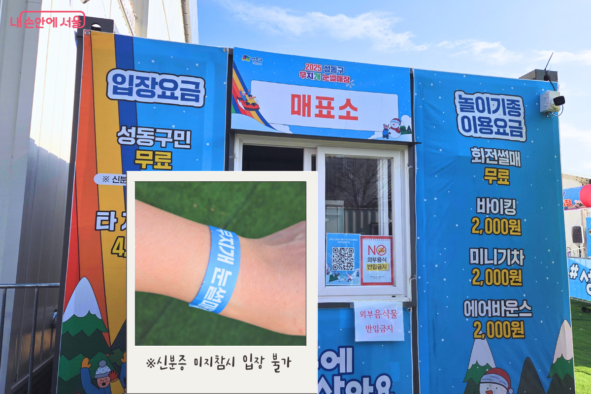파란색 배경의 현수막으로 꾸며진 눈썰매장 매표소의 모습이다. 왼쪽에는 '입장요금 성동구민 무료'라는 안내와 함께 신분증 미지참 시 입장이 불가하다는 경고 문구가 적혀 있고, 오른쪽에는 회전썰매 무료, 바이킹&middot;미니기차&middot;에어바운스 2,000원 등 놀이기구 이용 요금이 상세히 안내되어 있다. 사진 중앙에는 '무지개 눈썰매장' 문구가 적힌 파란색 입장 팔찌를 착용한 손목 모습이 합성되어 강조되어 있다. &copy;양정화