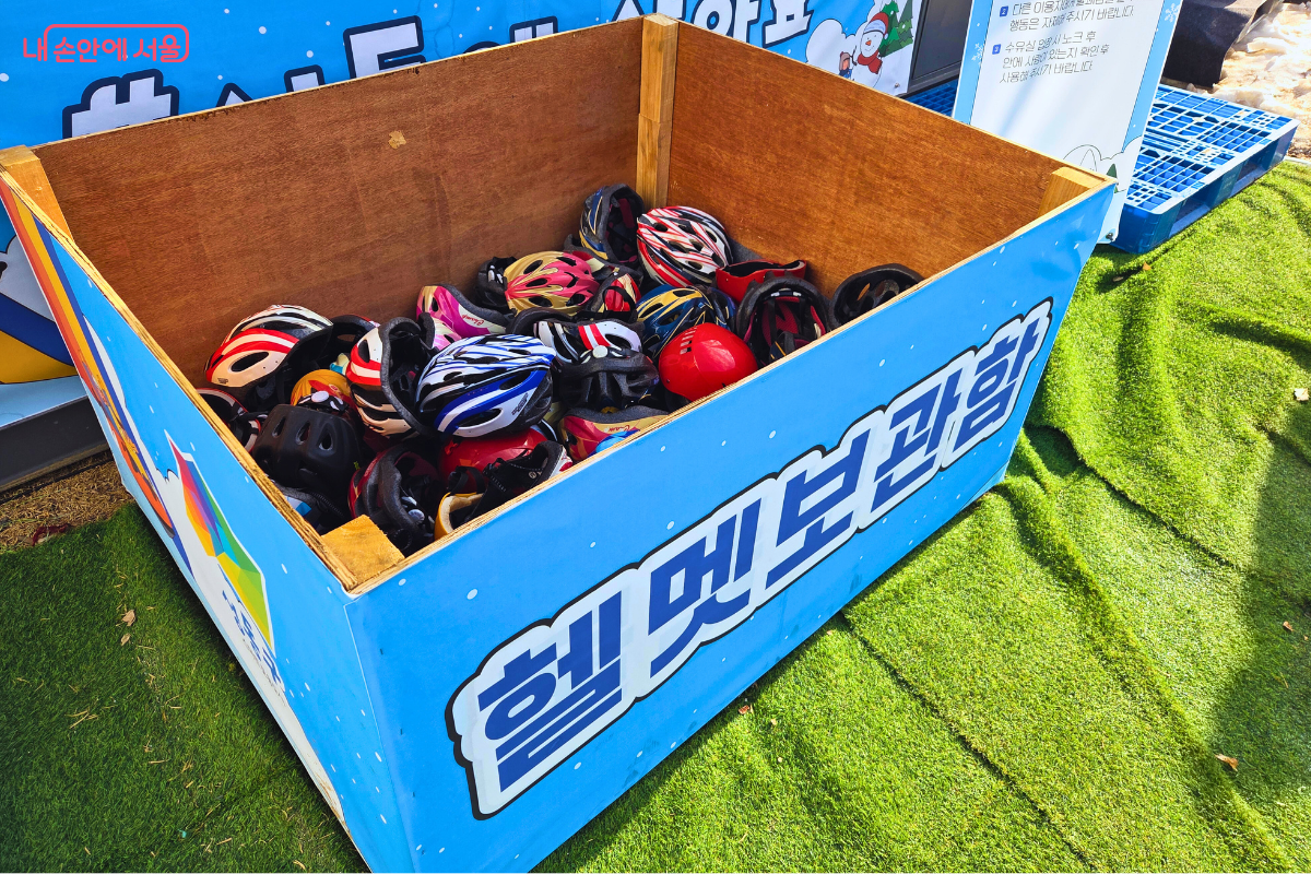 초록색 인조 잔디 위에 놓인 파란색 '헬멧보관함' 나무 상자 안에 다양한 색상과 디자인의 어린이용 헬멧이 가득 담겨 있는 모습이다. 파란색, 빨간색, 노란색 등 알록달록한 안전 헬멧들이 정리되어 있으며, 상자 뒤로는 시설 이용 안내판이 세워져 있어 이용객들의 안전 장구 착용을 독려하고 있다. &copy;양정화