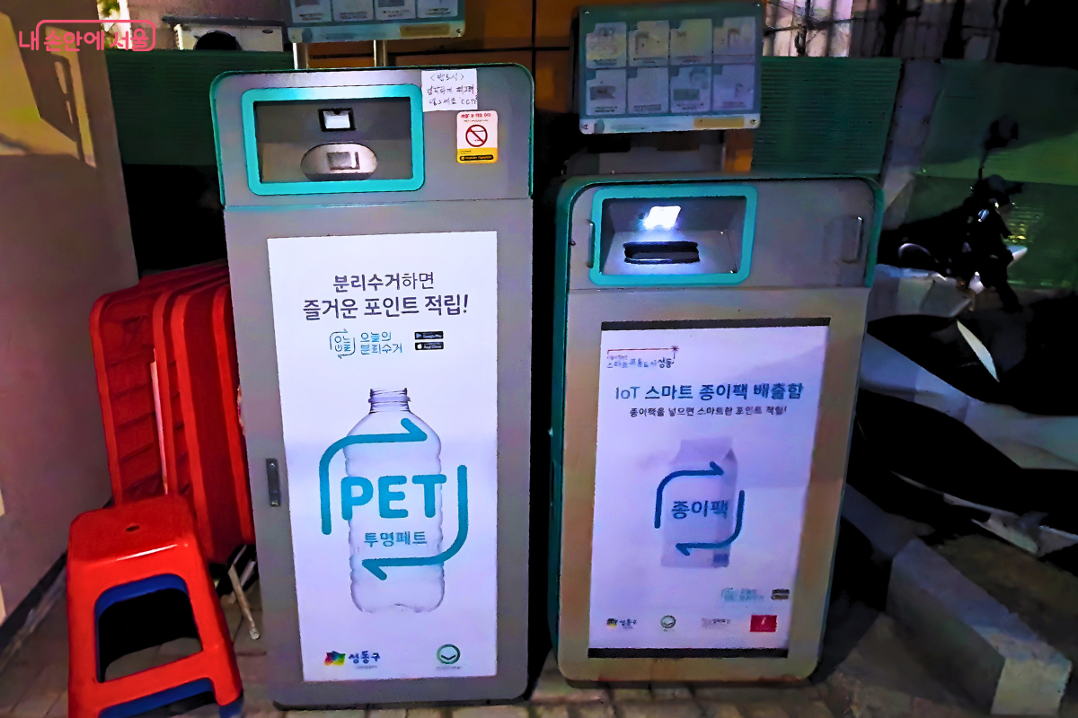 성동공유센터 외부에 나란히 설치된 두 대의 무인 회수기 모습이다. 왼쪽은 '투명 페트병(PET)' 전용 수거함이고 오른쪽은 '종이팩' 전용 수거함이다. 기기 전면의 안내판에 불이 들어와 있어 밤늦은 시간에도 시민들이 편리하게 분리배출을 할 수 있는 환경을 보여준다.