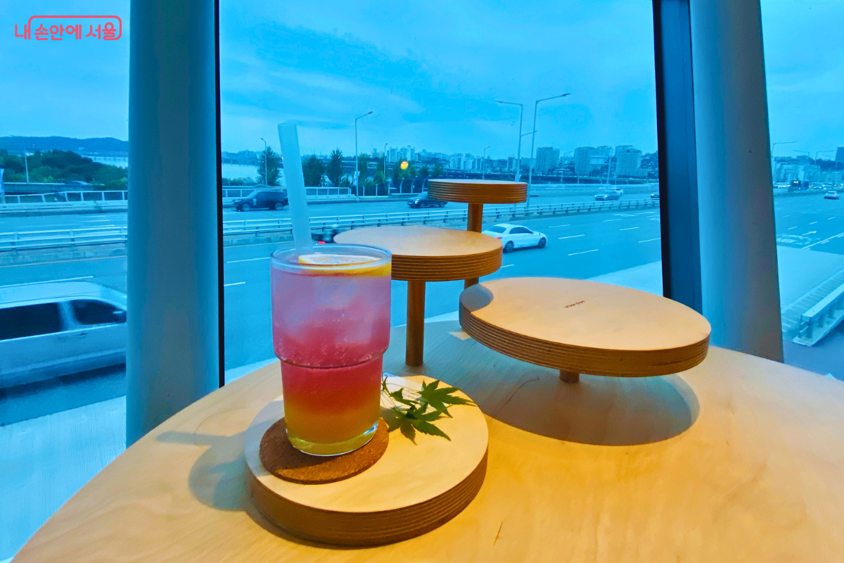 나무로 된 원형 스탠드 위에 놓인 노란색과 붉은색이 층을 이룬 에이드 음료의 모습이다. 유리잔 안에는 레몬 슬라이스가 들어있으며, 옆에는 작은 단풍잎 모양 식물이 장식되어 감성적인 분위기를 연출한다. &copy;양정화