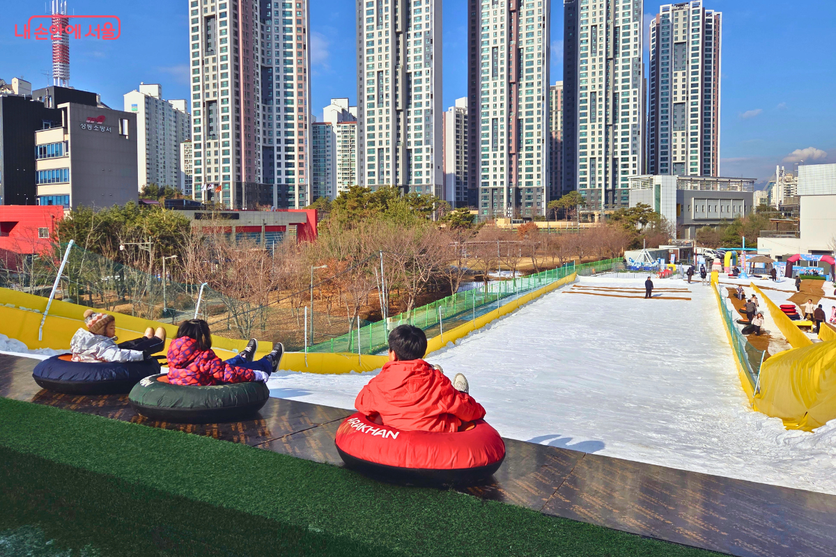 슬로프 정상에서 아래를 내려다본 대형 슬로프의 전체 모습이다. 신장 121cm 이상의 이용객만 사용할 수 있는 이 슬로프는 폭이 넓고 길게 아래로 뻗어 있으며, 출발 지점에 빨간색 점퍼를 입은 어린이가 튜브에 앉아 출발을 기다리고 있다. 슬로프 하단부에는 넓은 안전지대가 확보되어 있고, 저 멀리 다른 놀이기구들과 고층 빌딩이 어우러진 도심 전경이 한눈에 들어온다. &copy;양정화