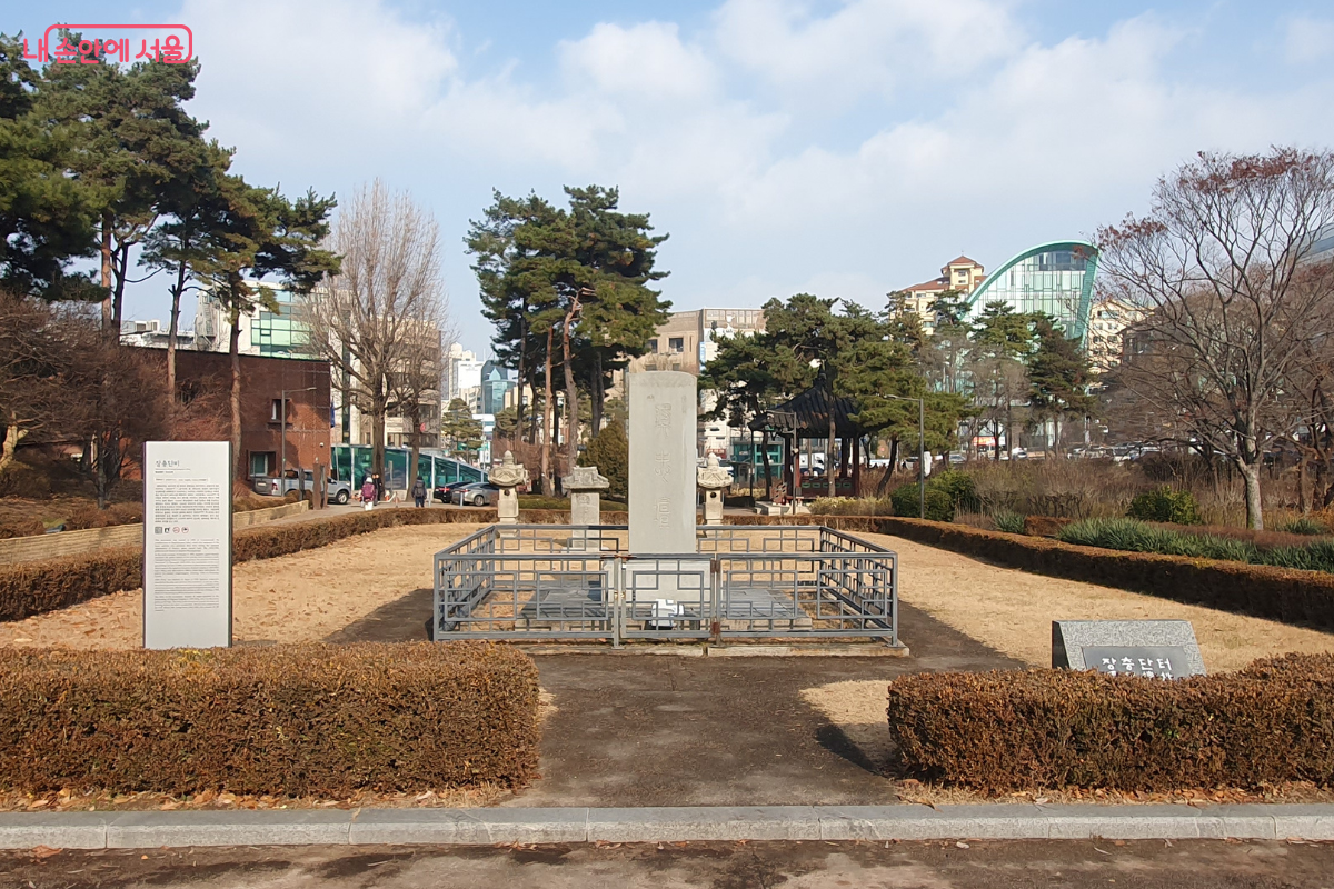 낮은 나무 울타리와 철제 난간으로 둘러싸인 장충단비의 전경이다. 중앙에 높게 솟은 석비가 있고 그 옆으로 작은 석등 형태의 구조물들이 배치되어 있다. 왼쪽에는 상세한 설명을 담은 안내판이, 오른쪽 앞쪽에는 '장충단 터'라고 적힌 표석이 놓여 있다. 뒤편으로는 소나무들과 현대식 건물들이 대조를 이룬다. &copy;양정화