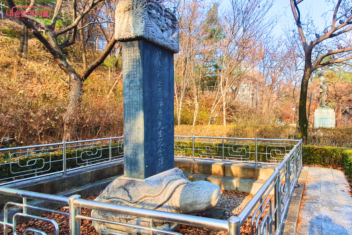 화강암으로 조각된 커다란 거북 받침대(비희) 위에 세워진 이한응 열사 기념비의 모습이다. 비석 상단에는 화려한 구름과 용 문양이 조각되어 있으며, 비문에는 열사의 공적이 기록되어 있다. 기념비 주변은 스테인리스 난간으로 보호되어 있고, 멀리 배경으로 이준 열사의 동상이 부분적으로 보인다. &copy;양정화