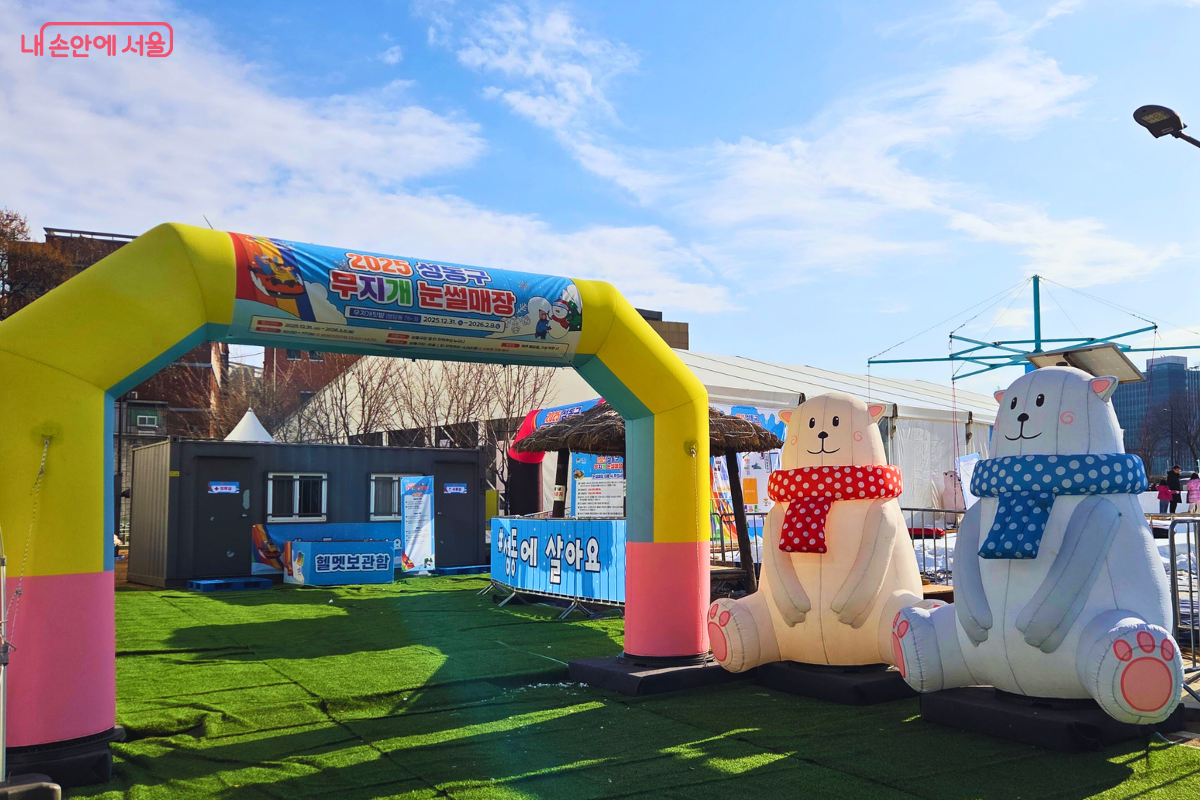 하얀 구름이 뜬 파란 하늘 아래 설치된 성동구 무지개 눈썰매장의 입구 아치 전경이다. 노란색과 분홍색 기둥으로 된 에어 아치에는 '2025 성동구 무지개 눈썰매장'이라는 문구가 적혀 있다. 입구 오른쪽에는 빨간색과 파란색 체크무늬 목도리를 두른 커다란 흰 곰 에어 인형 두 개가 나란히 앉아 방문객을 맞이하고 있으며, 바닥에는 초록색 인조 잔디가 깔려 있어 화사한 분위기를 자아낸다. &copy;양정화