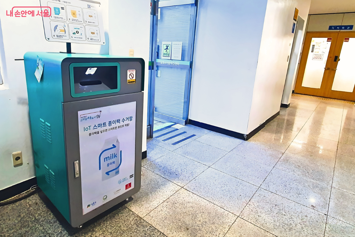 주민센터 내부 복도에 설치된 종이팩 수거함을 비스듬한 각도에서 촬영한 사진이다. 수거함은 자치회관 입구와 복도 창문 사이에 위치해 있어 시민들의 이동 동선에서 쉽게 발견할 수 있다. 