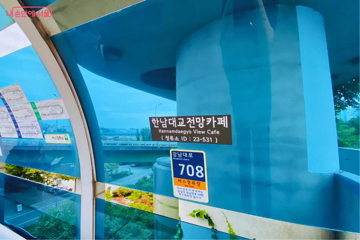 푸른색 유리 구조물에 '한남대교 전망카페(정류소 ID: 23-531)'라고 적힌 갈색 표지판이 부착되어 있고, 그 아래에는 건물의 도로명 주소인 '강남대로 708'이 명기된 안내 스티커가 붙어 있다. 유리창 너머로 한강 변 도로와 도시 풍경이 보이며, 왼편에는 버스 노선도가 부착되어 있어 카페와 정류장의 높은 접근성을 보여준다. &copy;양정화
