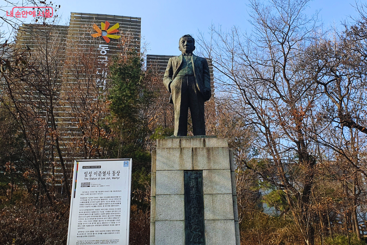 높은 화강암 기단 위에 세워진 일성 이준 열사의 전신 동상이다. 동상은 정장을 입고 당당하게 서 있는 모습이며, 기단 정면에는 열사의 이름이 한자로 새겨져 있다. 왼쪽에는 열사의 생애와 독립운동 업적을 기리는 안내판이 배치되어 있으며, 뒤편으로는 나무들 사이로 동국대학교 건물의 외관이 보인다. &copy;양정화