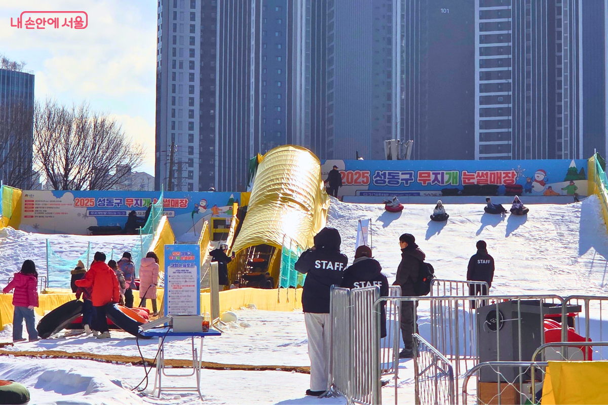 높이 솟은 고층 아파트 단지를 배경으로 펼쳐진 도심 속 눈썰매장의 전체 전경이다. 하얀 눈이 덮인 슬로프에서는 시민들이 튜브 썰매를 타고 내려오고 있으며, 슬로프 아래쪽에서는 등 뒤에 '스태프(STAFF)'라고 적힌 검은색 점퍼를 입은 안전요원들이 현장을 관리하고 있다. 노란색 천막으로 된 터널형 통로가 슬로프 위쪽까지 이어져 있으며, 입구 쪽에는 이용 수칙이 적힌 파란색 안내판이 세워져 있다. &copy;양정화