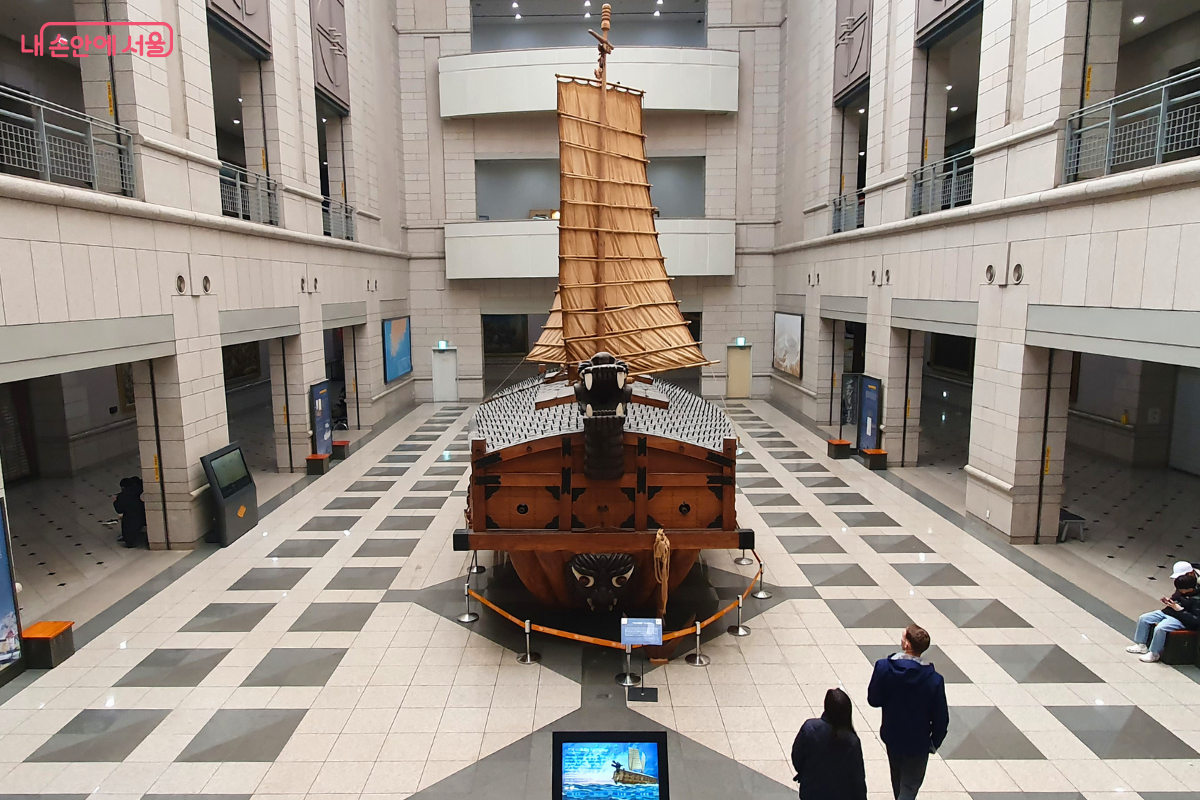 전쟁기념관 본관 내부 중앙 홀에 전시된 거북선 실물 모형을 위에서 내려다본 시점이다. 거북선은 용머리와 등에 박힌 송곳 등 세부적인 특징이 재현되어 있으며, 돛이 높게 펼쳐져 있다. 다층 구조의 실내 복도와 천장의 조명이 거북선을 비추고 있으며 관람객들이 주변을 둘러보고 있다.