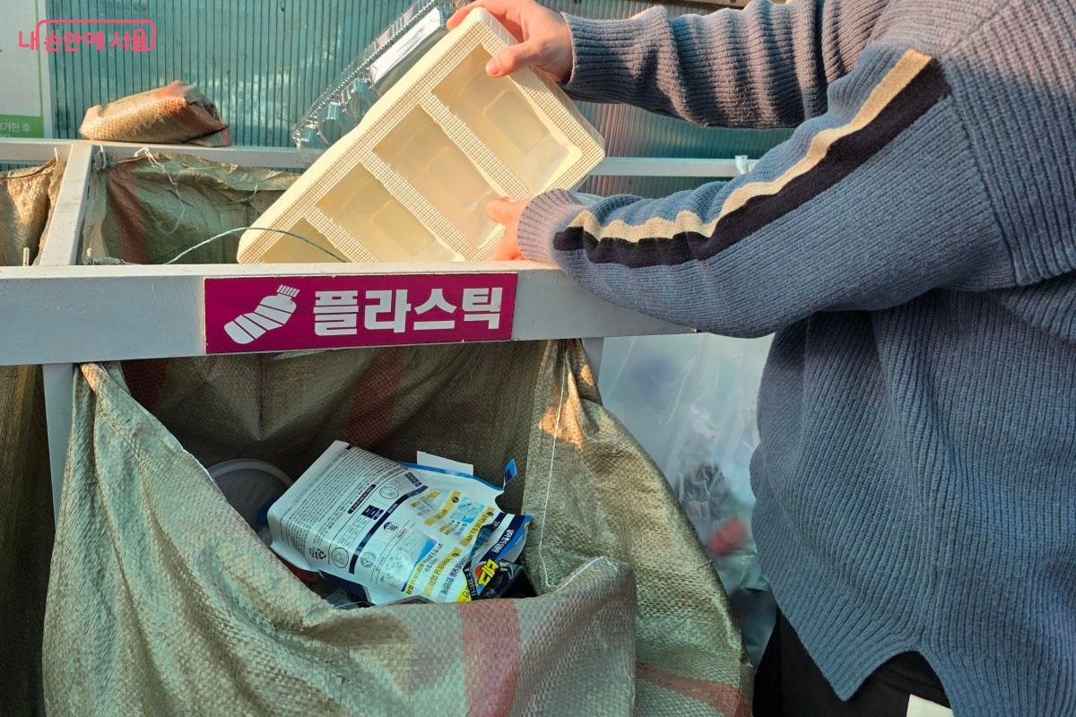 플라스틱 수거함에 배달 용기나 햇반 그릇 같은 플라스틱류를 차곡차곡 쌓아 분리배출하는 장면입니다.