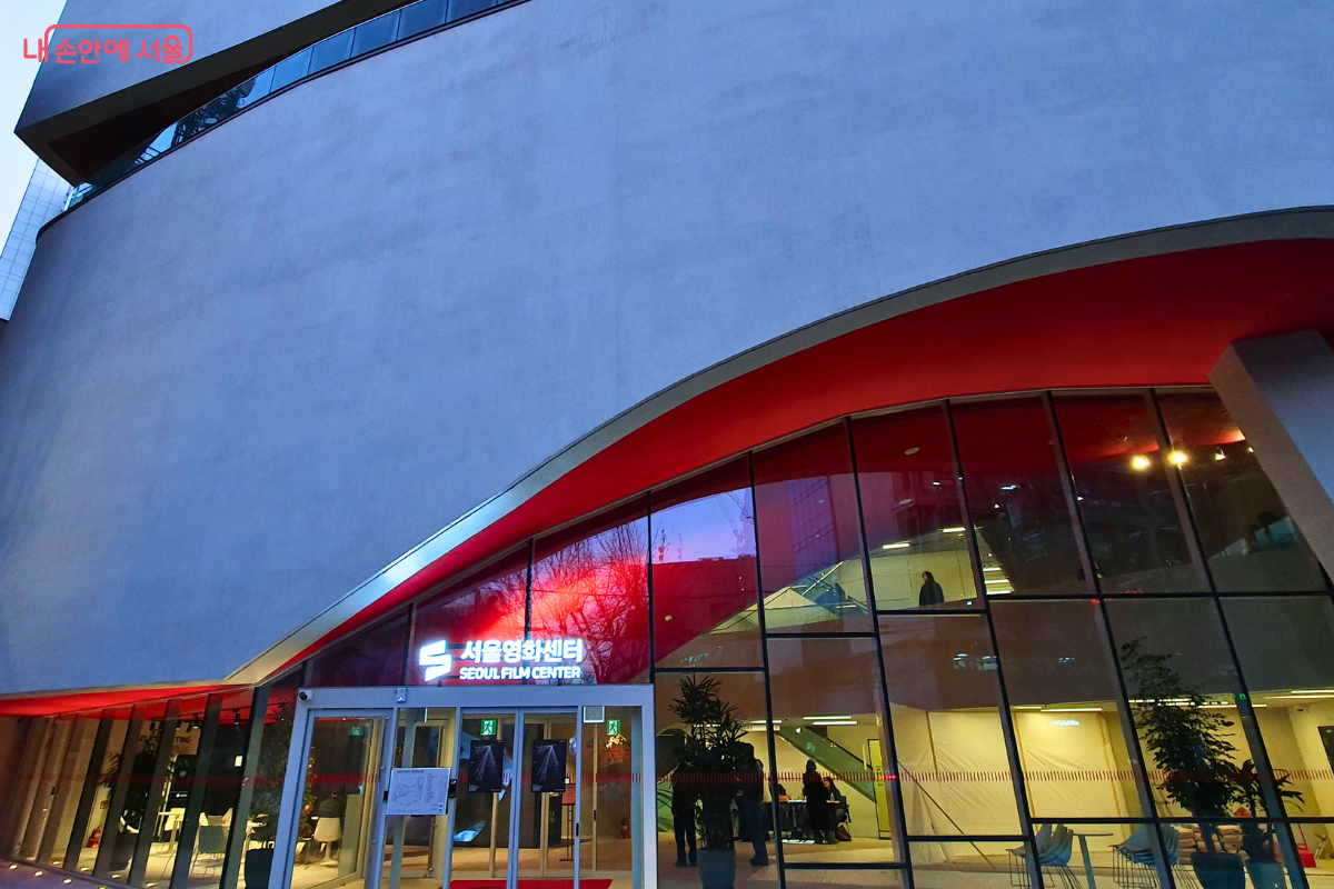 해질녘 서울영화센터 1층 유리 출입구를 가까이서 촬영한 사진이다. 사선으로 깎인 듯한 건물 하단부와 입구 천장의 강렬한 붉은색 포인트 조명이 세련된 대비를 이룬다. 투명한 유리창 안쪽으로는 밝은 조명의 로비와 안내 데스크, 방문객들의 실루엣이 보이며 문 위에는 '서울영화센터 SEOUL FILM CENTER'라는 공식 명칭이 하얀 글씨로 선명하게 빛나고 있다. ©양정화