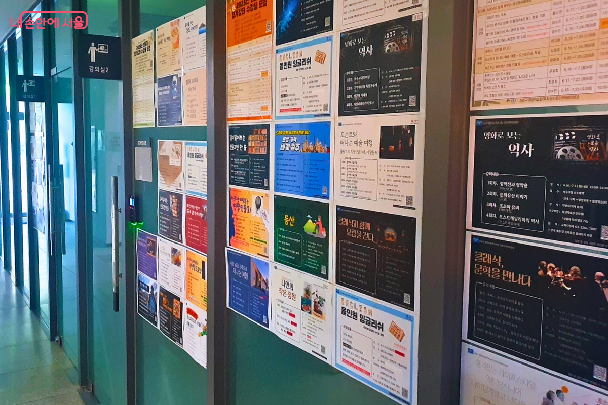 센터 내부 복도의 모습이다. 좌측에는 유리문으로 된 '강의실 1', '강의실 2' 입구가 보이며, 복도 벽면 전체에는 센터에서 운영 중인 각종 강좌와 특강 홍보 포스터들이 빼곡하게 부착되어 있다. 인문학, 예술, 외국어 등 다양한 교육이 이곳에서 이루어지고 있음을 짐작하게 하는 활기찬 교육 현장의 복도 풍경이다. &copy;양정화