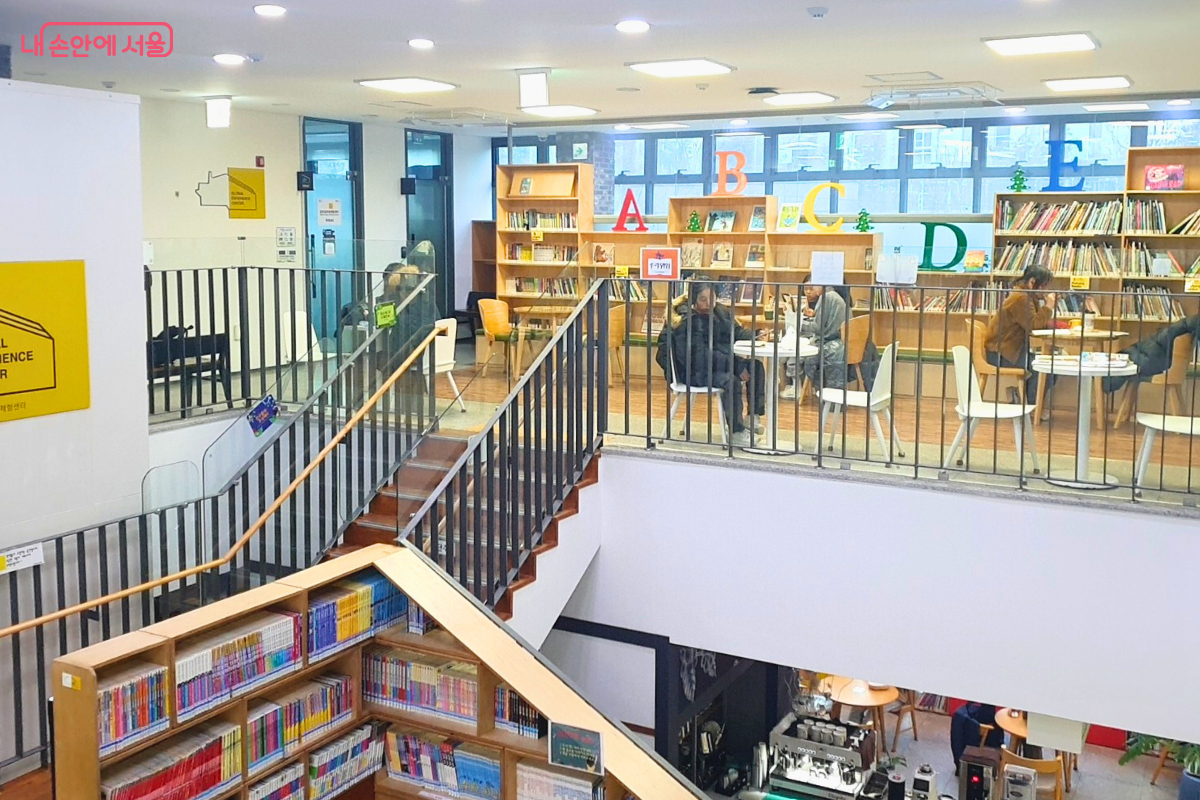 센터 2층에서 1층 책마루를 내려다본 시점의 사진이다. 2층 난간 옆으로는 'A, B, C' 등 알파벳 기호가 붙은 서가에 영문 서적들이 비치되어 있다. 중앙의 탁 트인 보이드 공간을 통해 1층의 카페와 서가가 내려다보이며, 창가 쪽에는 시민들이 앉아서 책을 보거나 대화를 나눌 수 있는 테이블 세트가 마련되어 있다. &copy;양정화