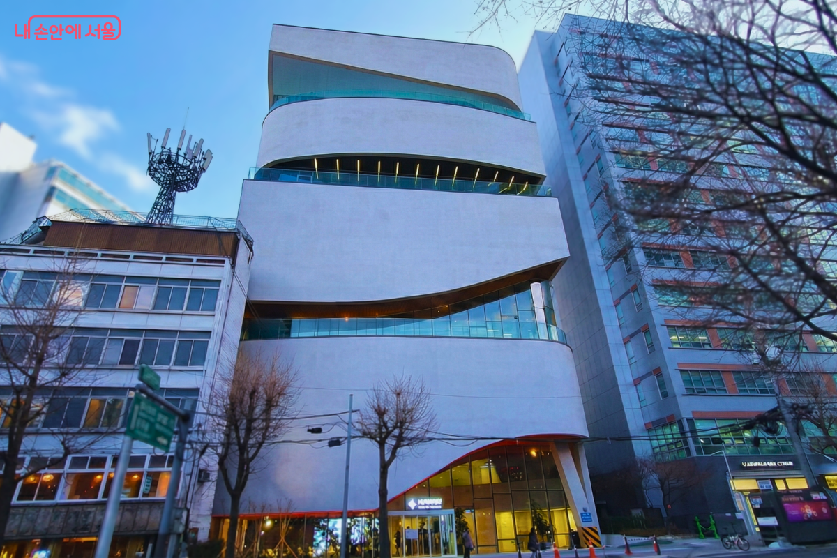 푸른 하늘을 배경으로 서 있는 서울영화센터의 건물 전체 모습이다. 흰색의 매끄러운 외벽이 층마다 비대칭적으로 겹쳐진 독특한 구조를 띠고 있으며, 건물 중간에는 가로로 길게 물결치는 모양의 유리창이 배치되어 현대적인 감각을 보여준다. 건물 주변으로는 잎이 떨어진 가로수들이 줄지어 서 있는 충무로 도심의 풍경이 담겨 있다. ©양정화