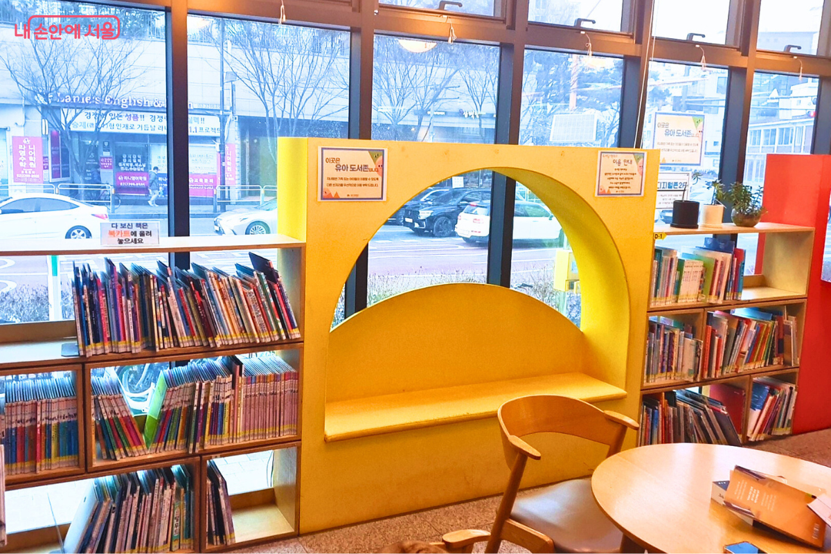 채광이 좋은 통유리창 옆에 조성된 유아 도서 존의 모습이다. 노란색으로 포인트를 준 아치형 벽면 좌석은 아이들이 쏙 들어가 책을 읽기에 안성맞춤이다. 서가에는 '이곳은 유아 도서존입니다'라는 안내문이 붙어 있으며, 창밖으로 도로 건너편의 풍경이 보여 시원한 개방감을 준다. 원형 테이블 위에는 읽다 만 책들이 놓여 있다. &copy;양정화