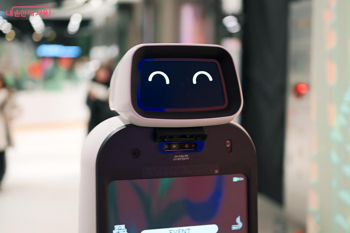 갤러리 내부에는, 최신식 이동형 스마트 AI로봇이 웃음짓는 표정을 지으며 돌아다니고 있다.