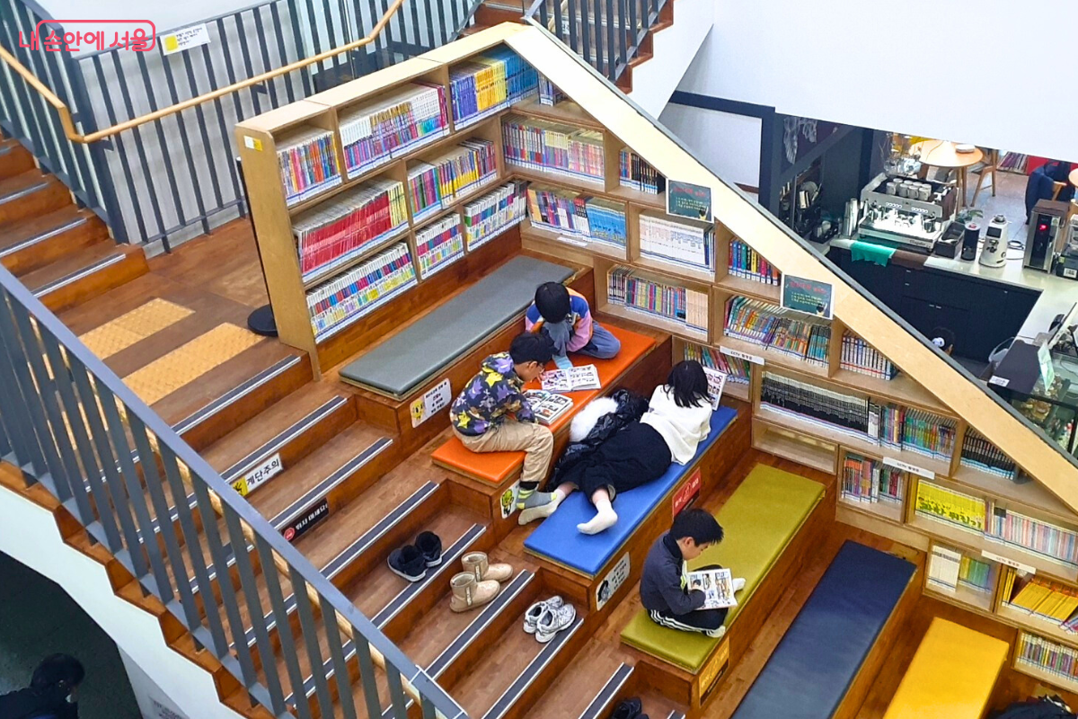 계단식으로 조성된 어린이 전용 독서 공간의 모습이다. 나무 소재로 만들어진 계단형 좌석 위에 어린이들이 신발을 벗고 올라가 편안한 자세로 책을 읽고 있다. 각 층에는 알록달록한 쿠션이 놓여 있으며, 계단 옆면을 활용한 서가에는 어린이용 전집과 만화책 등이 가득 꽂혀 있어 자유로운 독서 분위기를 조성한다. &copy;양정화