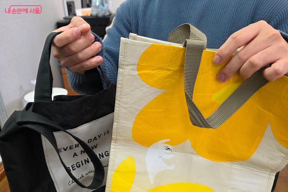 외출 시 비닐봉지 대신 사용하는 노란색 꽃무늬 에코백과 검은색 천 가방을 양손으로 들어 보이고 있습니다.