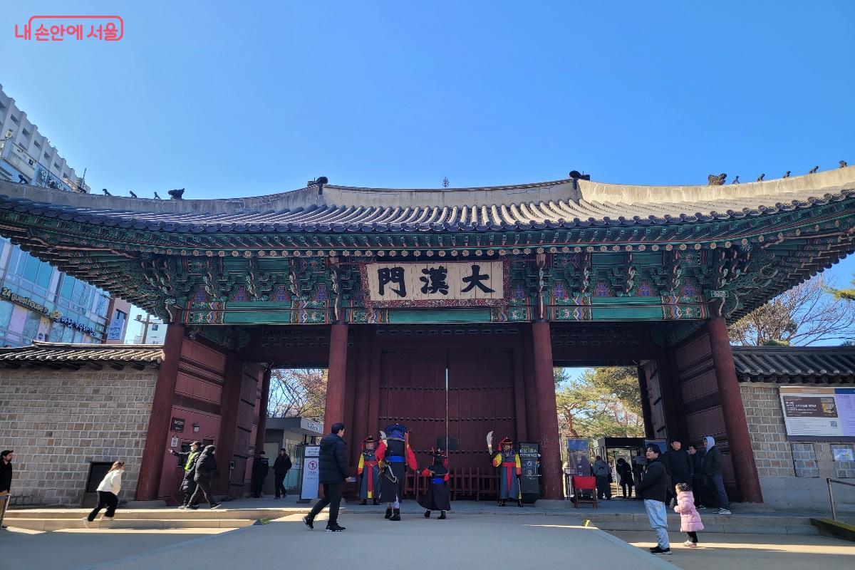 서울 덕수궁 대한문 전경으로 전통 단청이 돋보이는 궁궐 정문 앞에 수문장 복장의 인물들과 관람객들이 서 있는 모습.